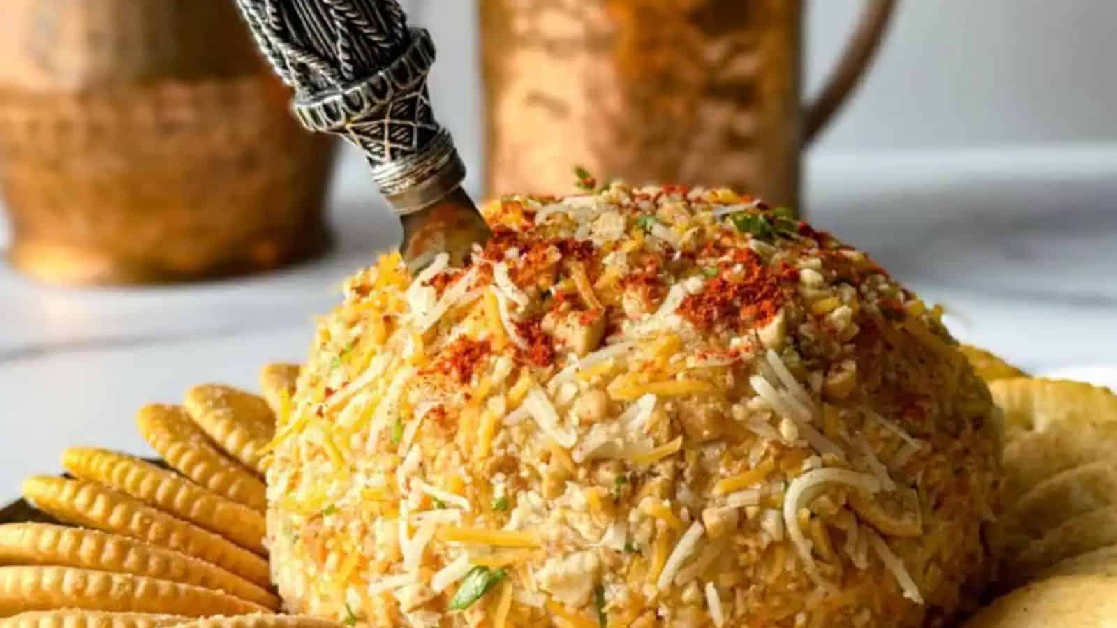 A cheese ball topped with spices and herbs is placed on a plate surrounded by crackers. An ornate knife is inserted into the top. Copper vessels are blurred in the background.