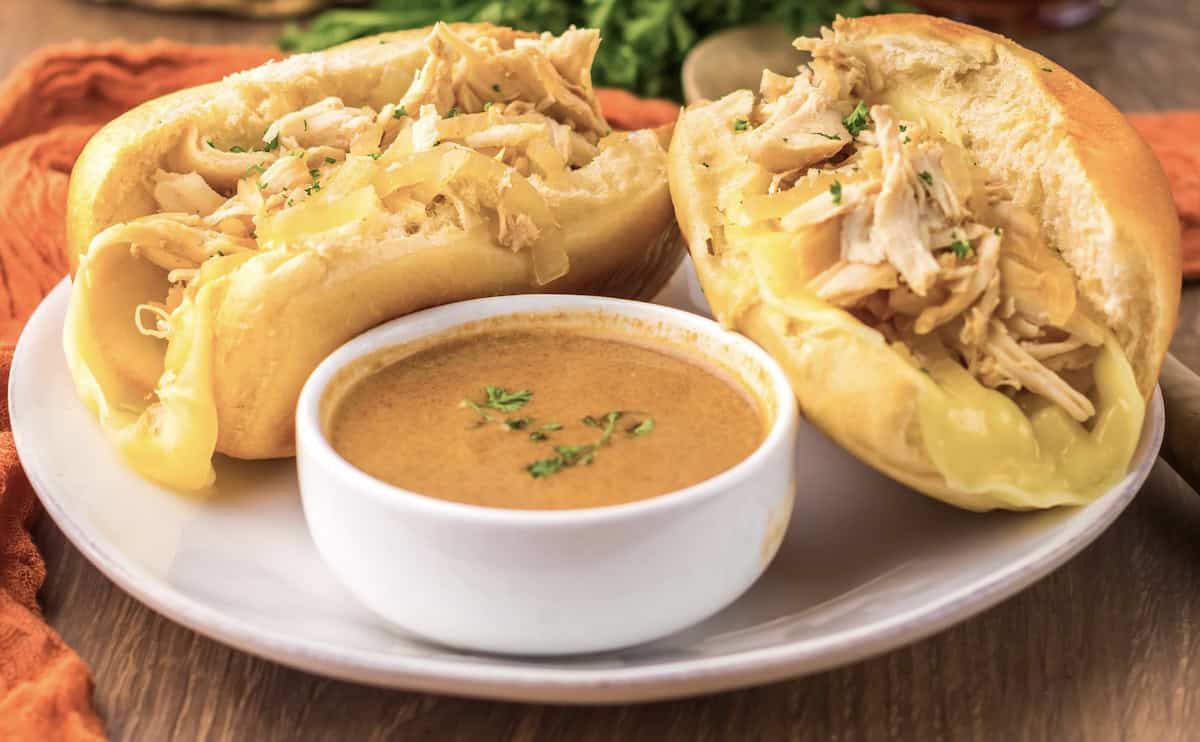 Two open-faced sandwiches with shredded chicken and melted cheese on soft rolls served with a side of dipping sauce in a small white bowl on a plate.