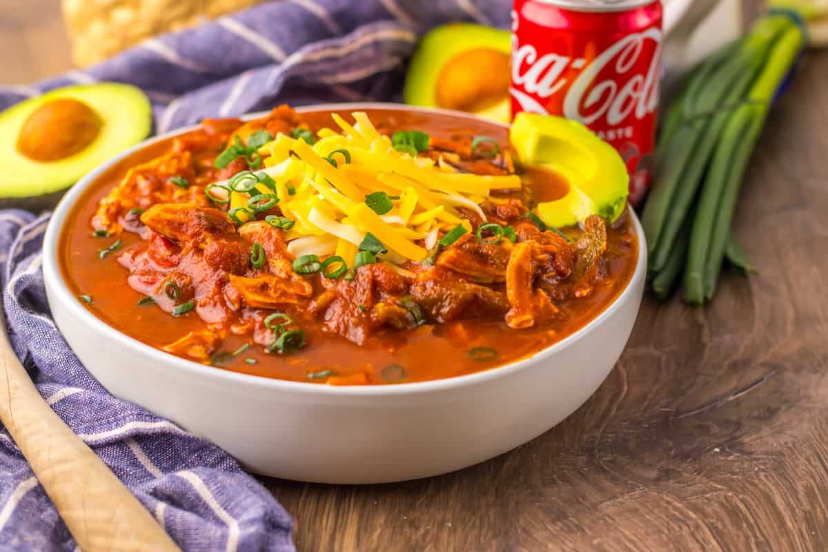 A bowl of chili topped with shredded cheese and green onions, accompanied by avocado slices. A soda can, whole avocado, and green onions are in the background on a wooden surface.