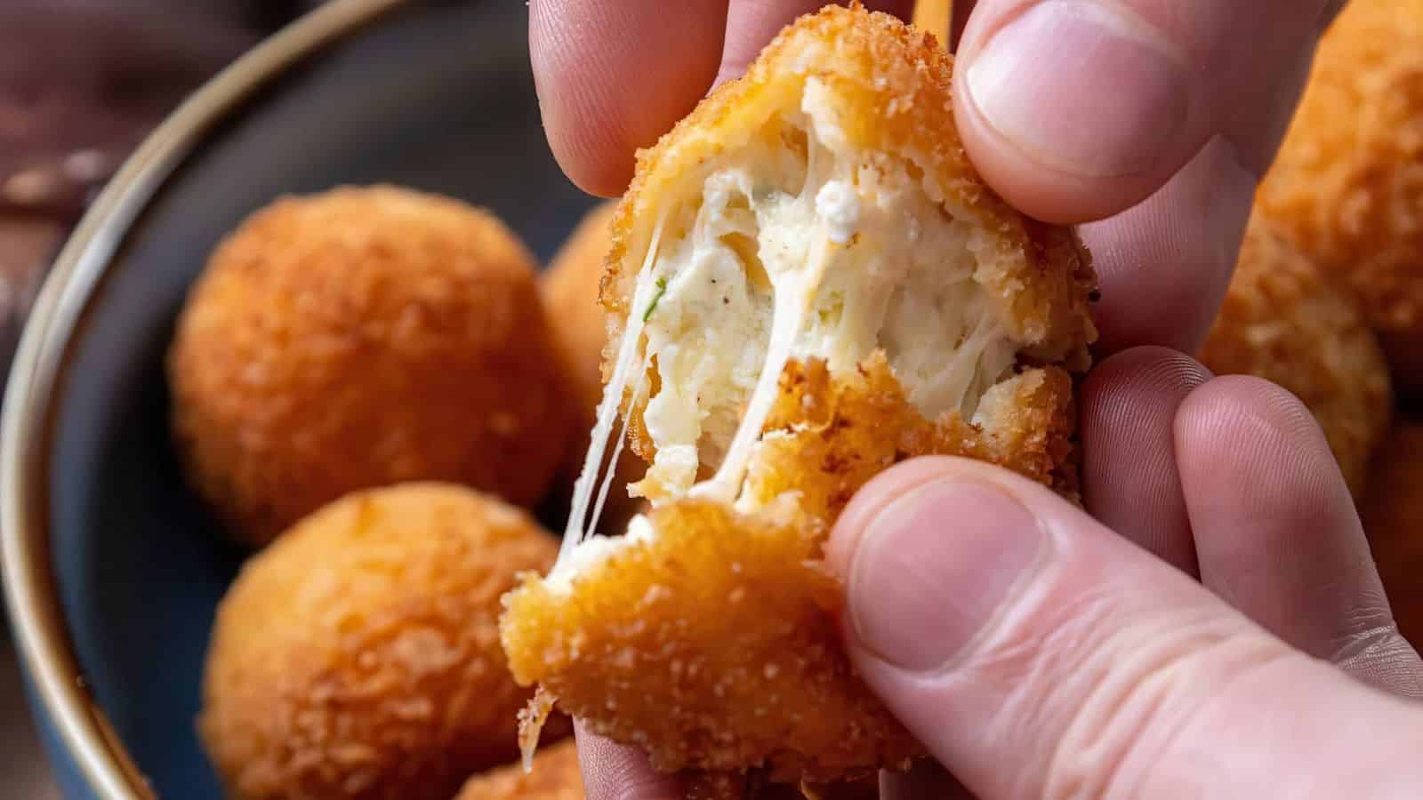 Close-up of hands pulling apart a crispy, golden-brown, fried cheese ball, revealing melted cheese inside. More cheese balls are visible in a bowl in the background.
