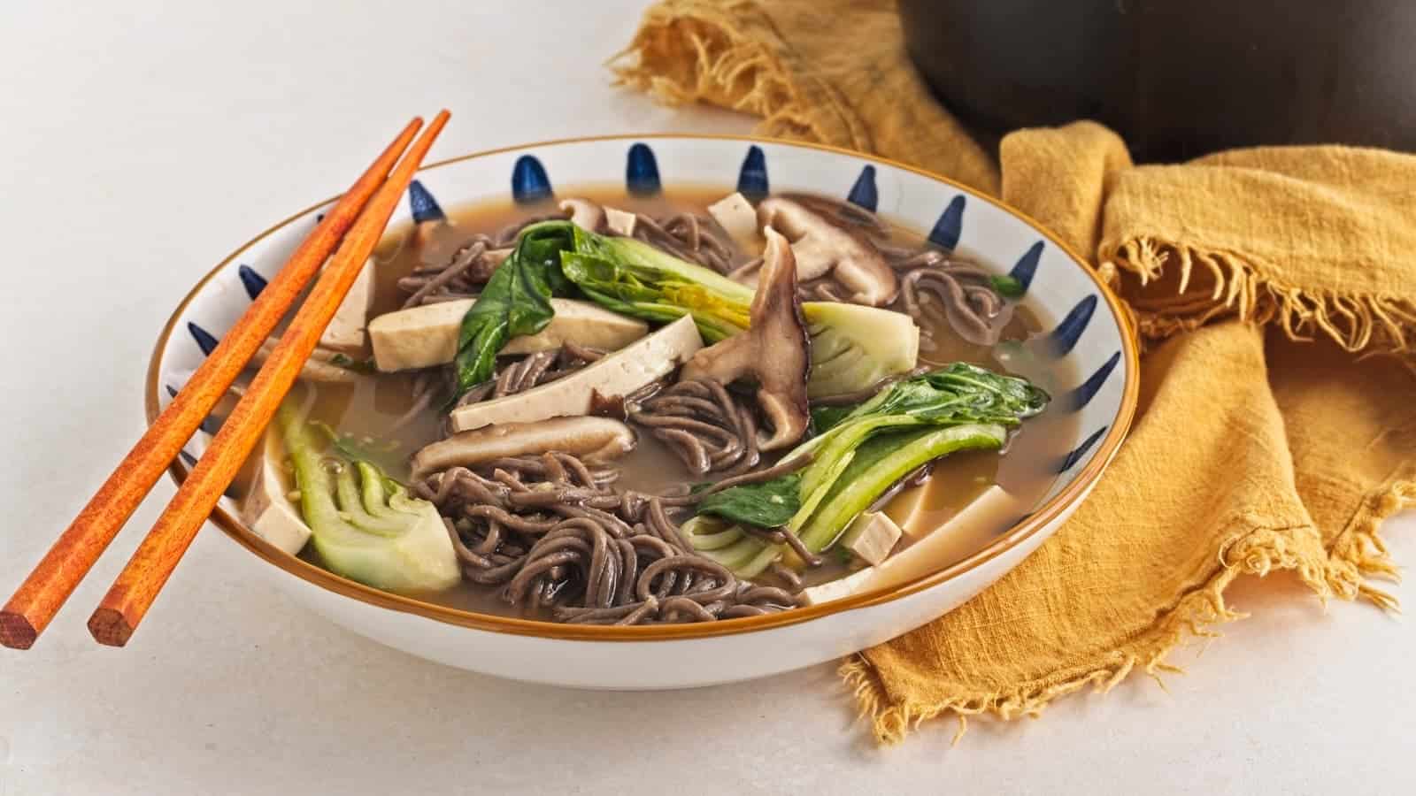 A bowl of soba noodle soup with mushrooms, tofu, and bok choy. Orange chopsticks rest on the bowl, and a yellow cloth is nearby.