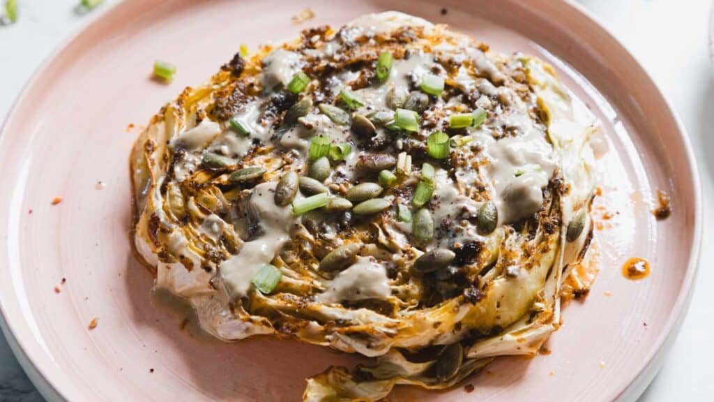 Roasted cabbage steak on a pink plate, topped with creamy sauce, seeds, and chopped green onions.