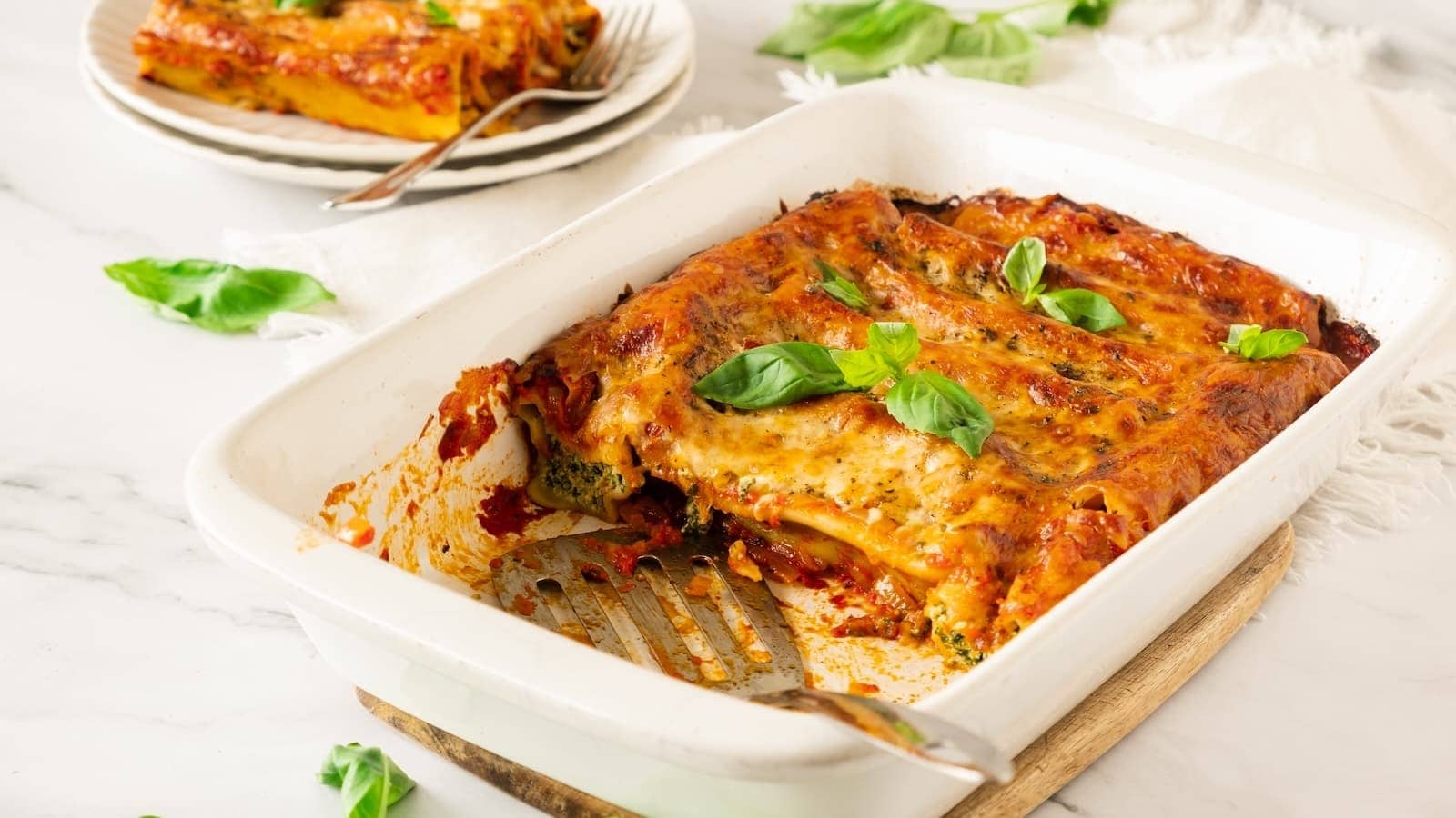 Baked spinach cannelloni in a white dish with a serving spatula, garnished with basil leaves. Plates with more servings and basil scattered around.