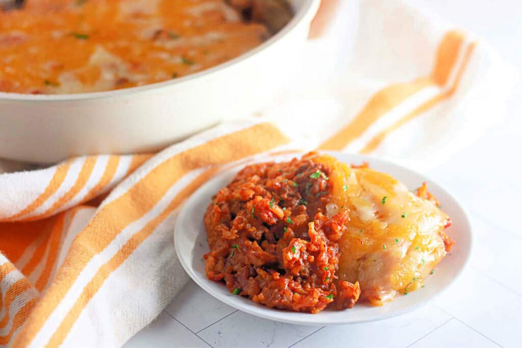 A small white plate with a serving of casserole topped with golden cheese and a portion of saucy, ground meat. A pot with more casserole is in the background, next to a striped towel.