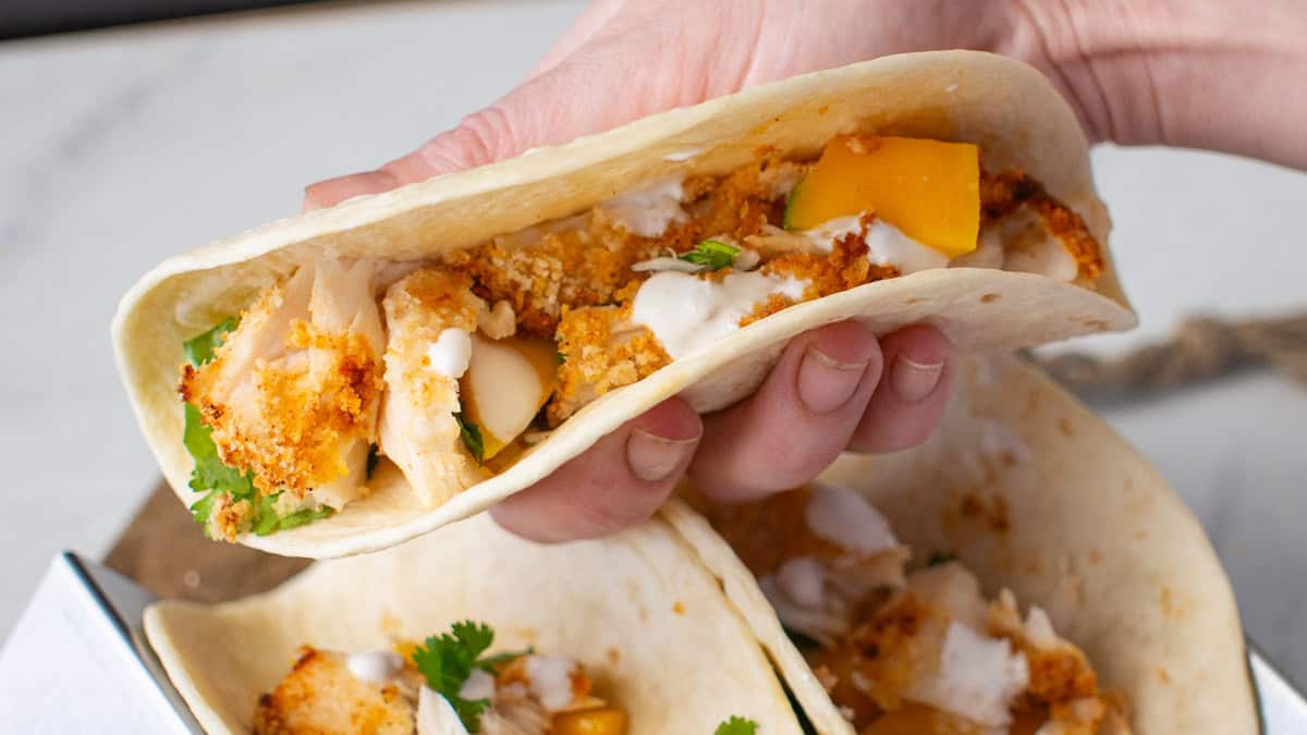 A hand holding a soft taco filled with breaded fish, diced mango, cilantro, and white sauce.