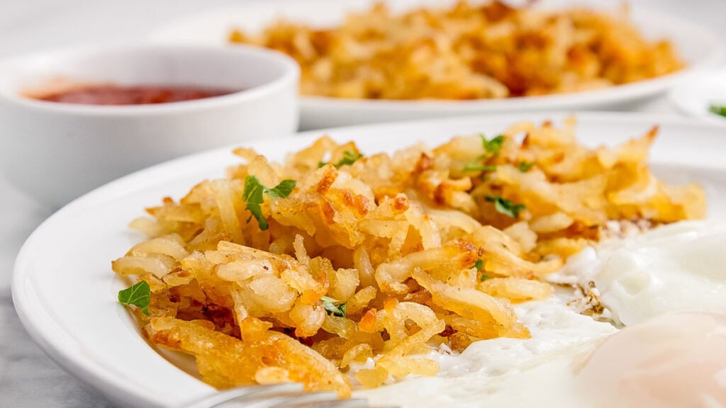 Close-up of a plate with hash browns and fried eggs. A small dish of red sauce and another plate of hash browns are in the background.