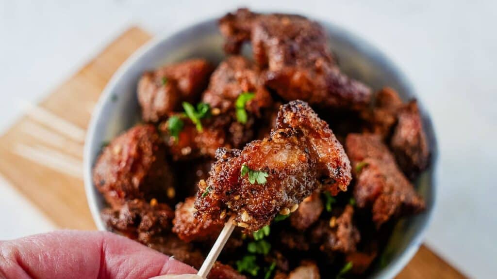 Close-up of a hand holding a toothpick with a piece of spiced fried meat. A bowl filled with similar pieces garnished with herbs is in the background.