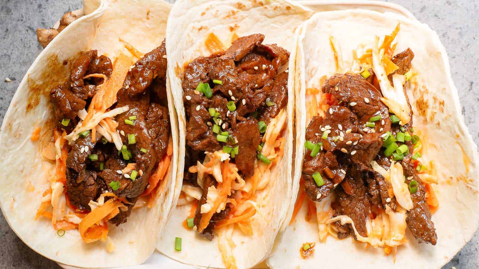 Horizontal shot of 3 korean tacos: corn tortillas filled with sliced steak, kimchi and pico de gallo salsa, and chopped cilantro. In the background are lime wedges, sliced red cabbage, and cilantro leaves.