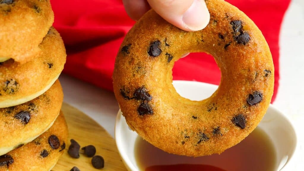 Hand holding a chocolate chip donut above a bowl of syrup; stack of donuts and scattered chips in the background.