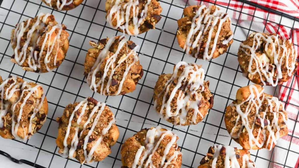 Muffins with crumb topping and icing drizzle on a cooling rack, next to a red and white cloth.