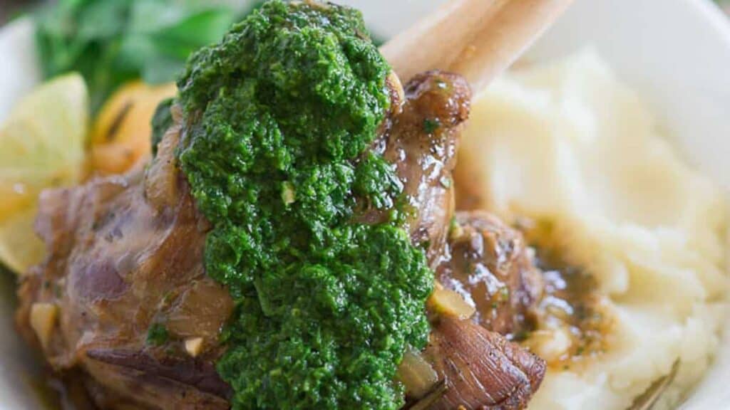 Lamb shank topped with green sauce served on a bed of mashed potatoes, with lemon wedges and spinach in the background.
