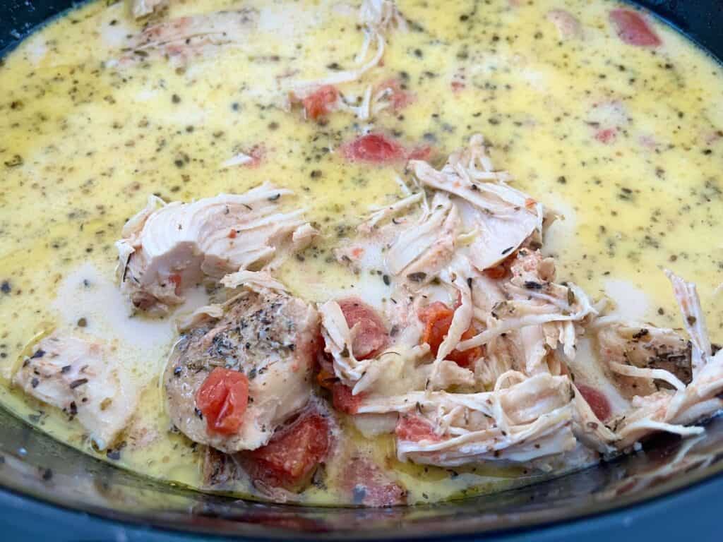 A slow cooker filled with a creamy soup containing shredded chicken, diced tomatoes, and herbs.