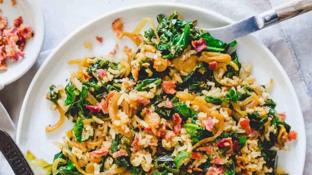 A plate of cooked greens with rice, topped with small bacon bits, is shown with a fork resting on the edge.
