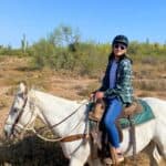 A girl rides a white horse through the desert landscape of Scottsdale wearing a helmet, sunglasses, plaid shirt, and jeans.