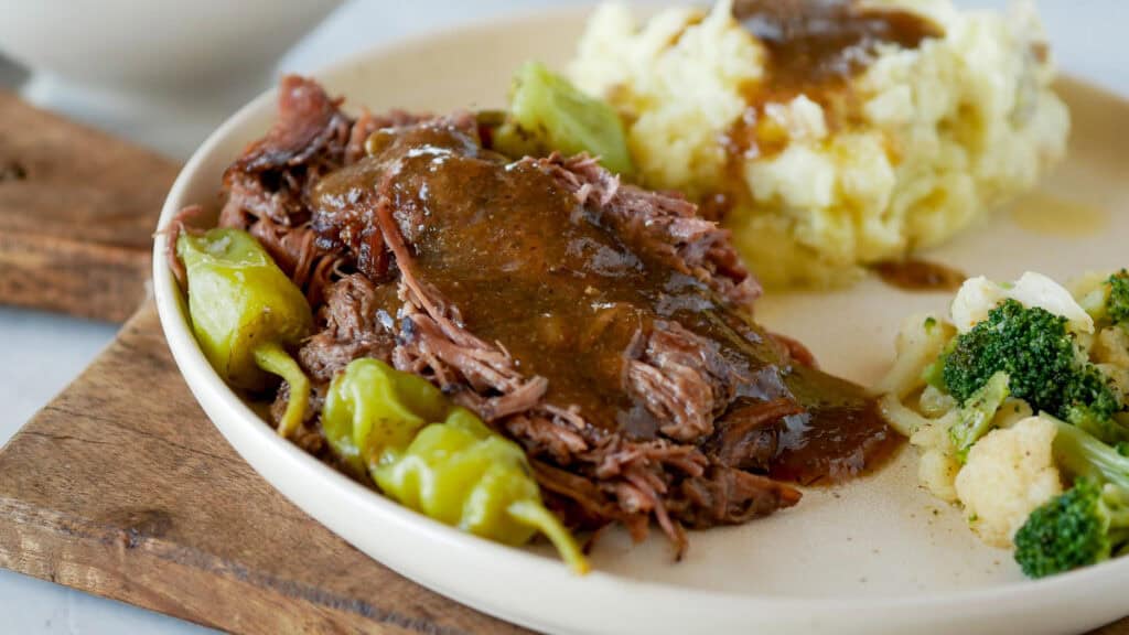Plate with shredded beef in gravy, pepperoncini, mashed potatoes, and broccoli with cauliflower on a wooden board.