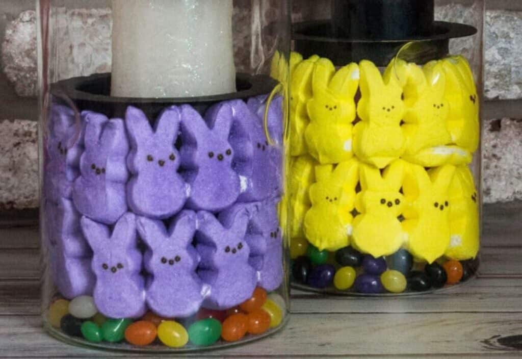 Glass jars filled with purple and yellow marshmallow bunny candies, much like peeps, surround white candles. Colorful jelly beans are layered at the bottom.