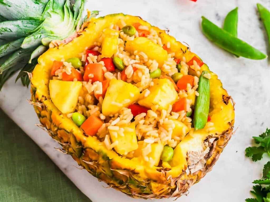 Pineapple half filled with rice, carrots, peas, and pineapple chunks, garnished with a green bean, on a light surface with green accents.