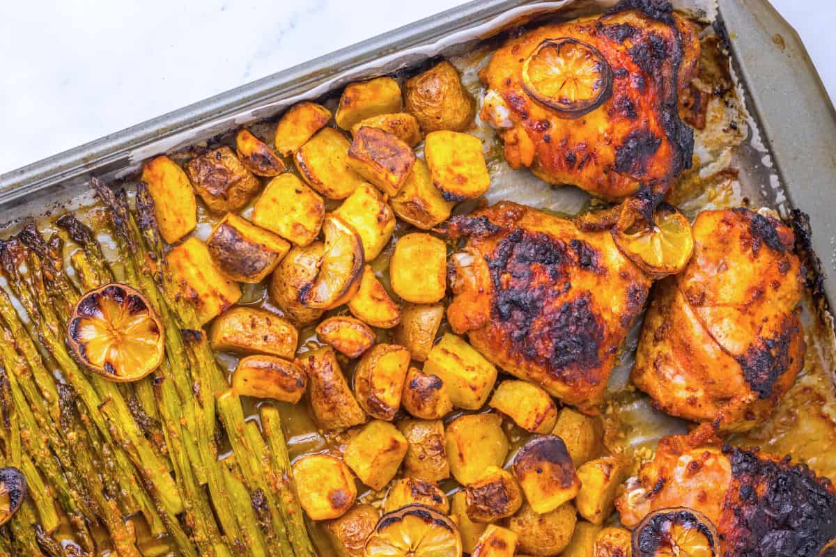 Baked chicken thighs, roasted potatoes, and asparagus with lemon slices on a baking sheet.