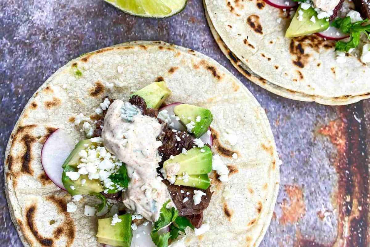 Three beef tacos with avocado, radish slices, cilantro, and crumbled cheese in taco holders on a textured surface.