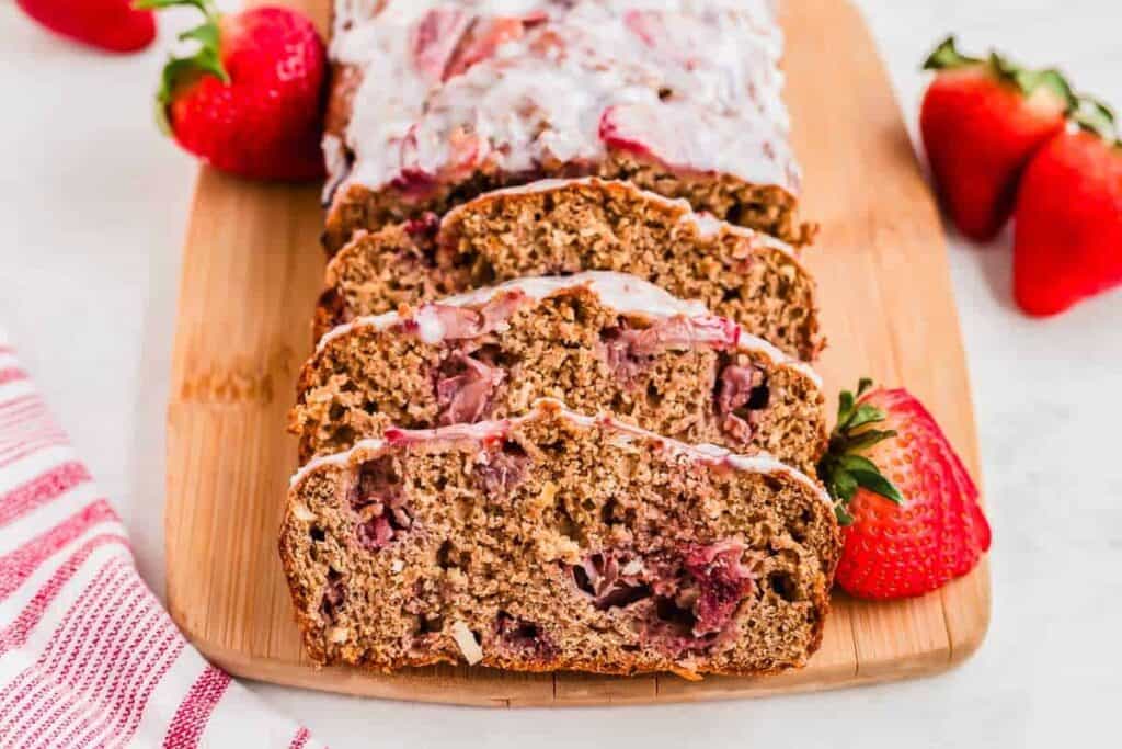 Sliced strawberry bread on a wooden board, surrounded by fresh strawberries. A red and white striped cloth is partially visible on the left.