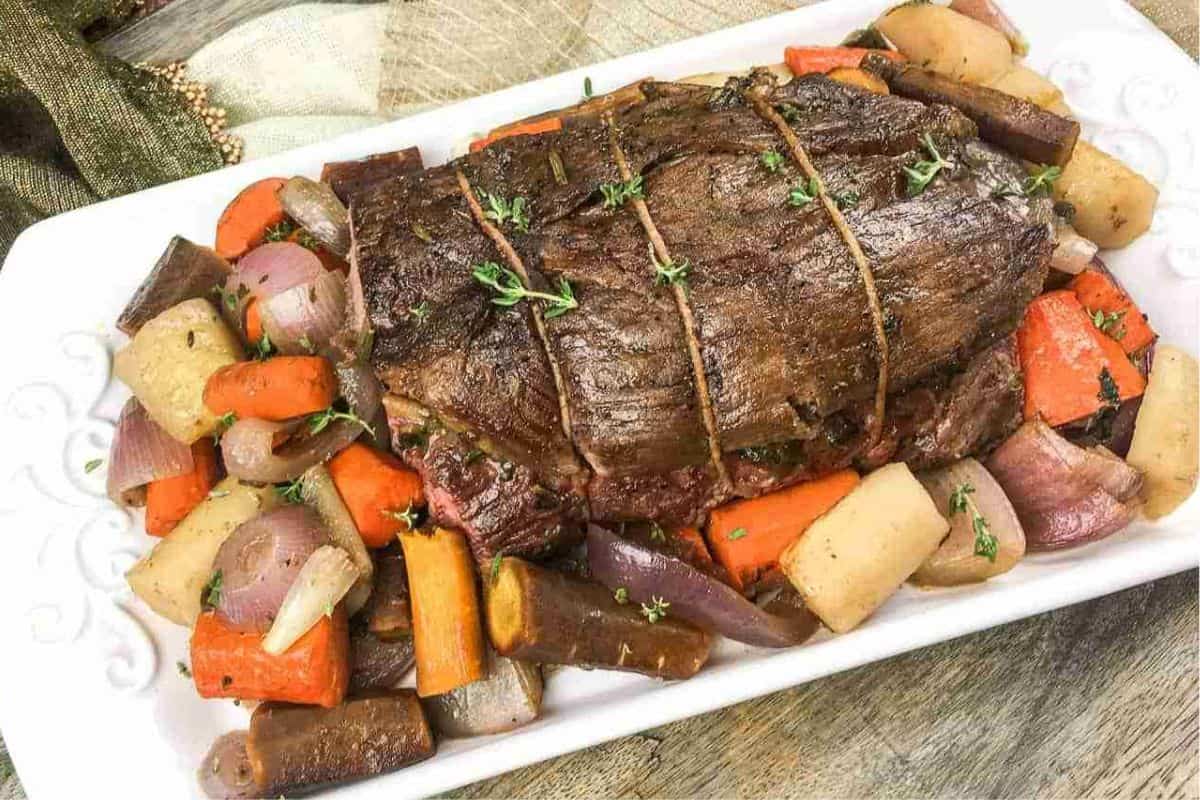 Roast beef tied with twine on a white platter, surrounded by roasted carrots, potatoes, and red onions.