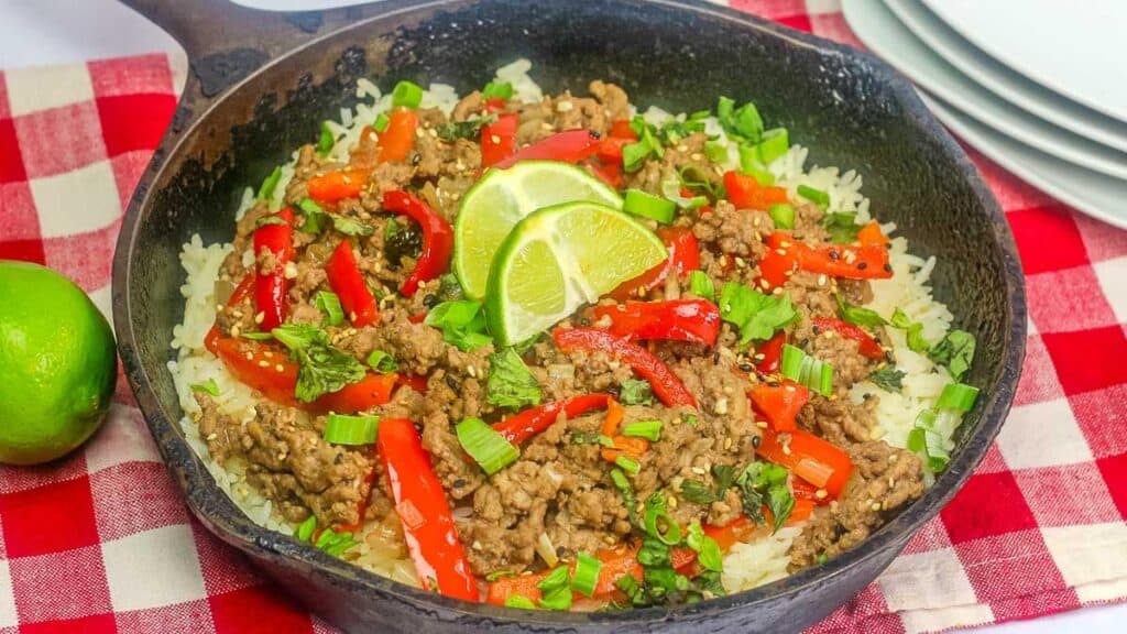Skillet with ground meat, red peppers, and green onions on rice, garnished with lime slices, on a red checkered cloth.
