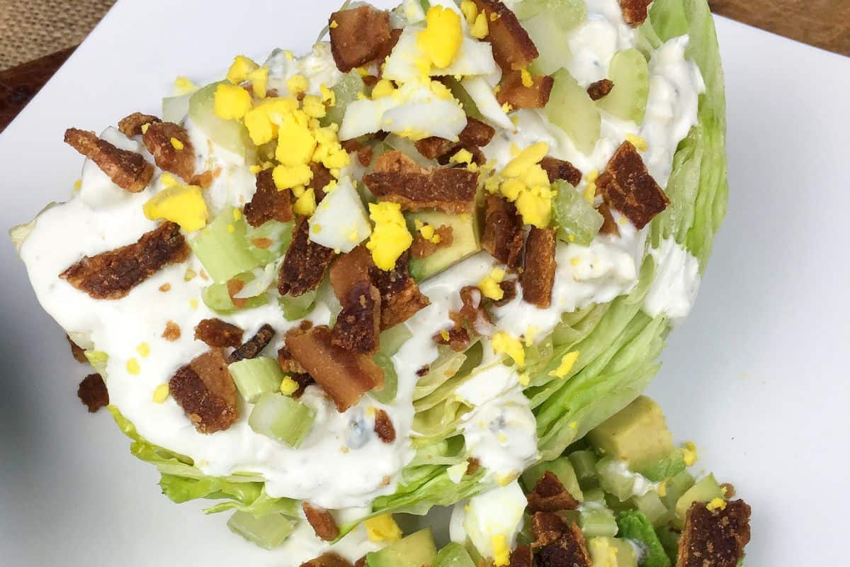 A wedge salad topped with crumbled bacon, chopped boiled egg, and creamy dressing on a white plate.
