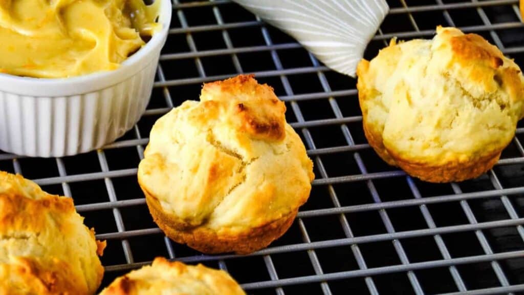 Three golden-brown biscuits rest on a cooling rack beside a white ramekin filled with creamy spread.