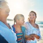 Three people, likely of different generations, enjoy a sunny beach walk together&mdash;smiling and laughing with waves and blue sky behind them&mdash;capturing the joy of multigenerational travel on a perfect grandymoon.