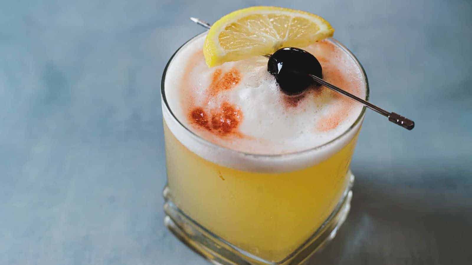 A cocktail in a glass, topped with a layer of frothy foam, garnished with a lemon wedge and a cherry on a skewer. The drink appears to be a whiskey sour, presented against a blurred blue-gray background.