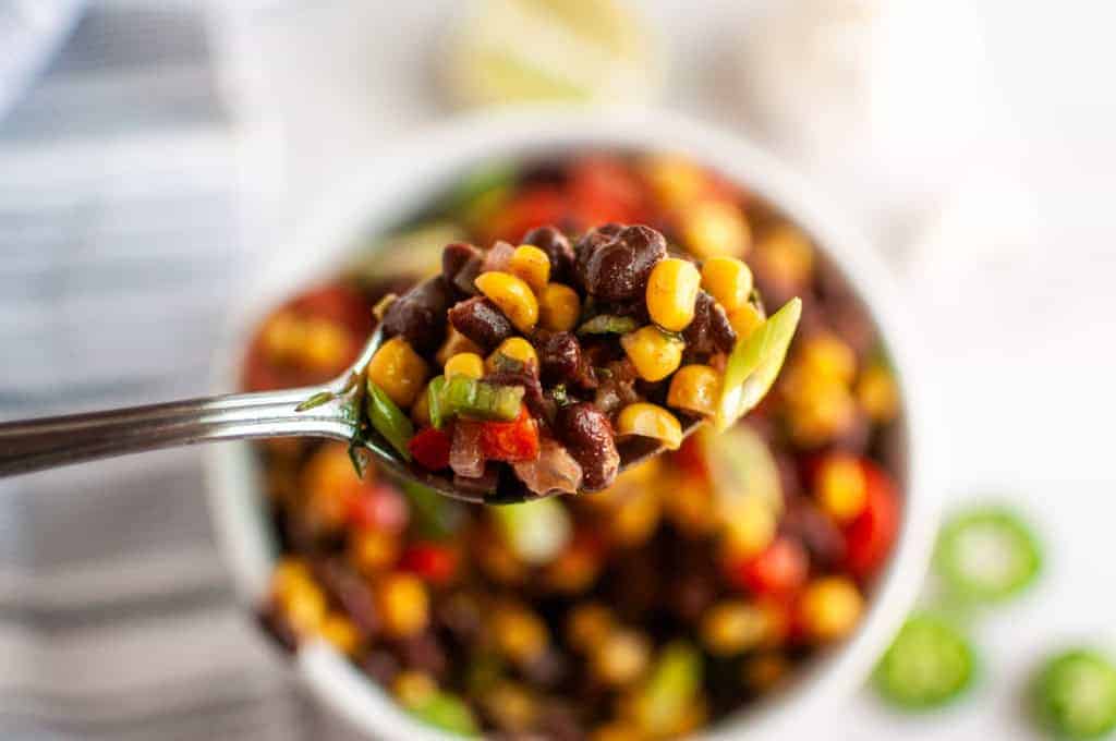 A close-up of a spoonful of black bean, corn, and vegetable salad held above a bowl filled with the same salad.