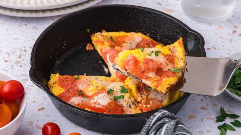 A slice of baked frittata with melted cheese, tomatoes, and herbs is lifted from a cast iron skillet with a spatula.
