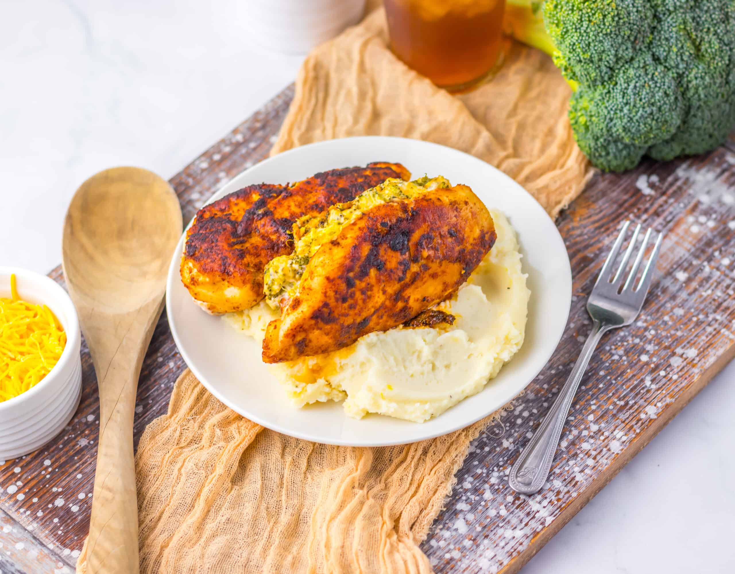 Plate of stuffed chicken breasts on mashed potatoes, with a wooden spoon and fork, next to a bowl of shredded cheese and broccoli on a wooden board.