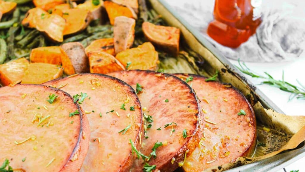 A white plate with a serving of glazed ham garnished with rosemary, surrounded by roasted sweet potato chunks and green beans. A gray napkin and a bottle of syrup are beside the plate.