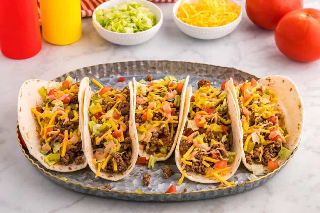 Five beef tacos with lettuce, cheese, and diced tomatoes on a metal tray. Bowls of shredded lettuce and cheese in the background. Ketchup and mustard bottles are visible.