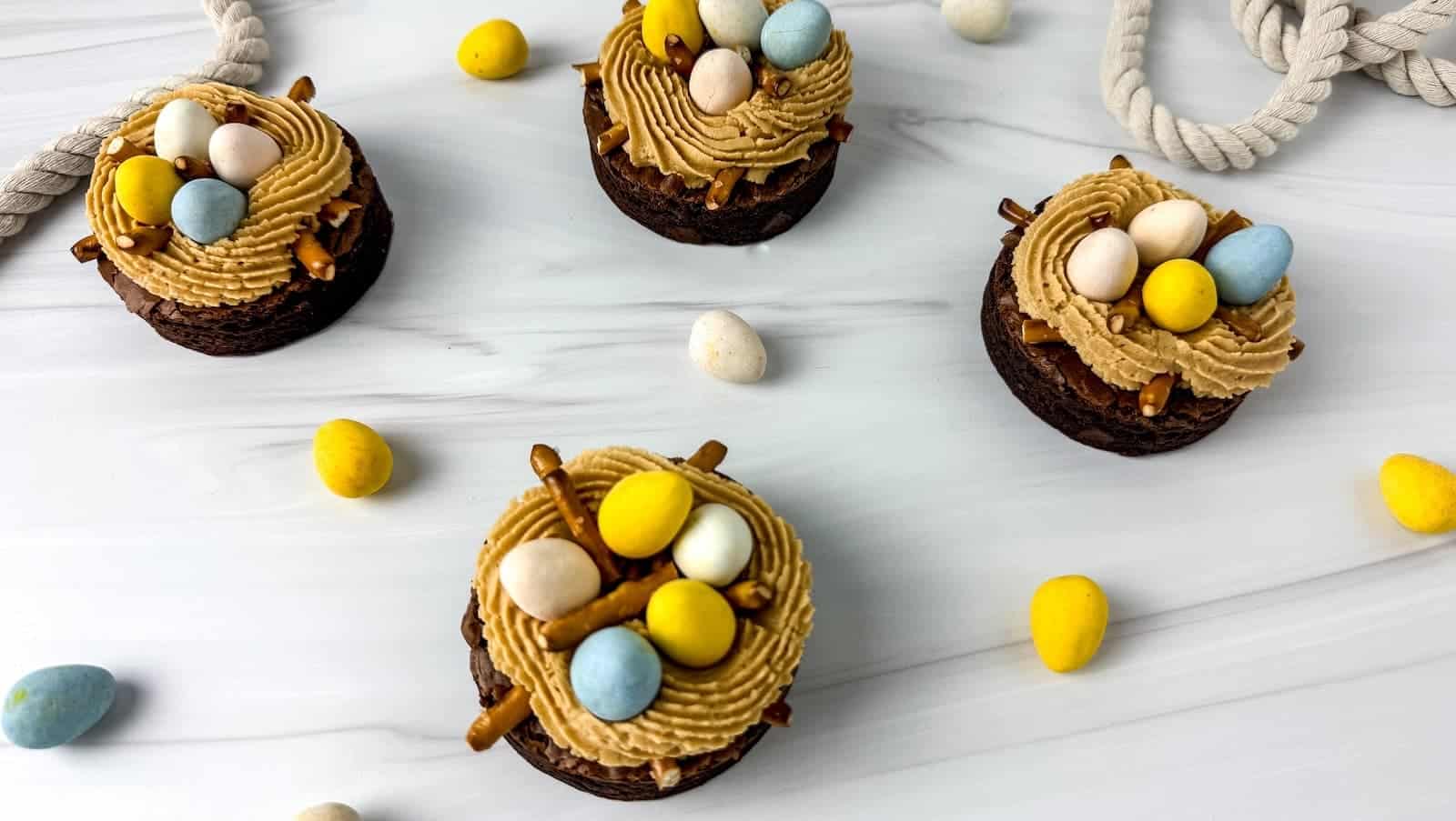 Four Easter brownies with brown frosting and colorful chocolate eggs sit on a white surface, surrounded by scattered candies and a gray rope.