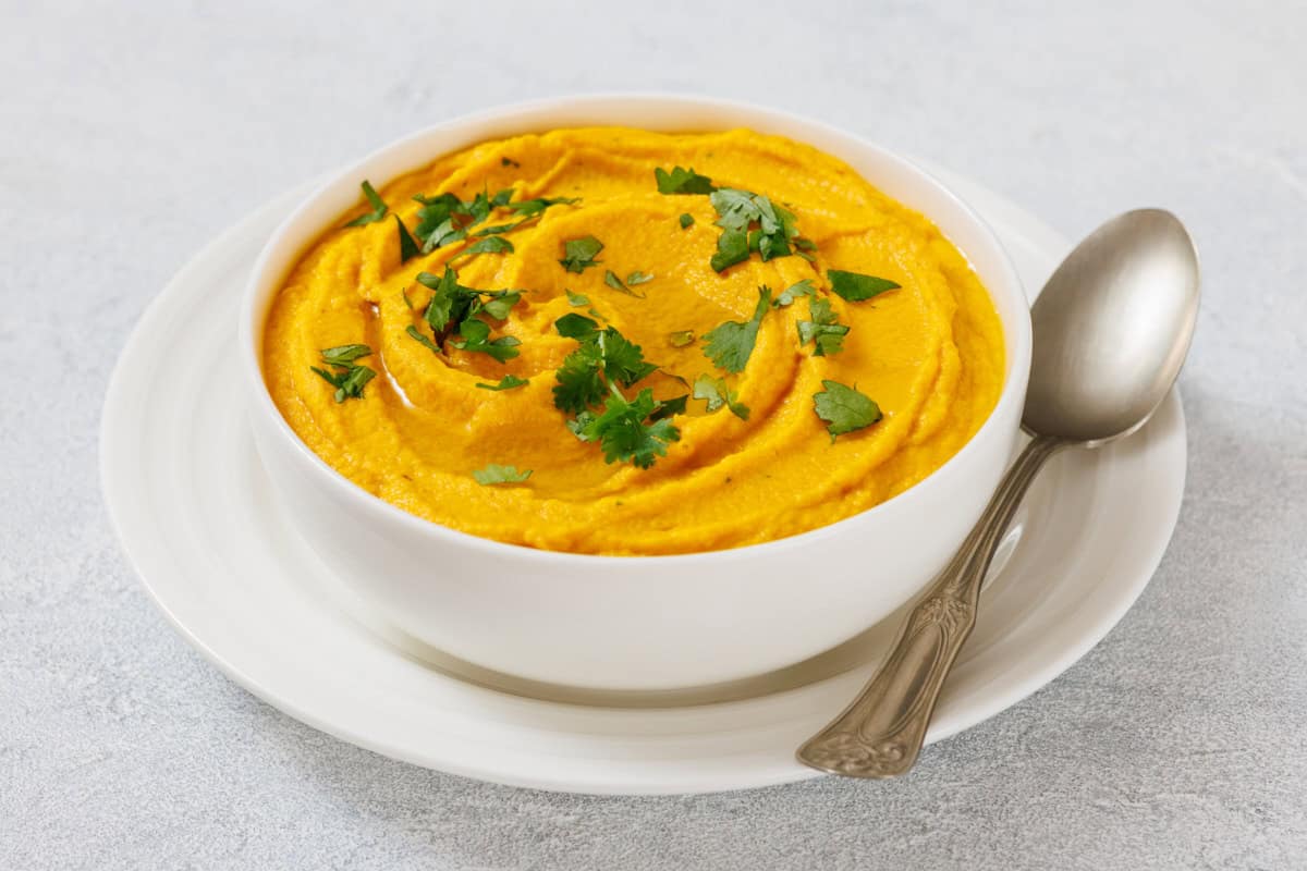 A bowl of Roasted Carrot Hummus garnished with chopped cilantro, placed on a white plate with a silver spoon on the side.