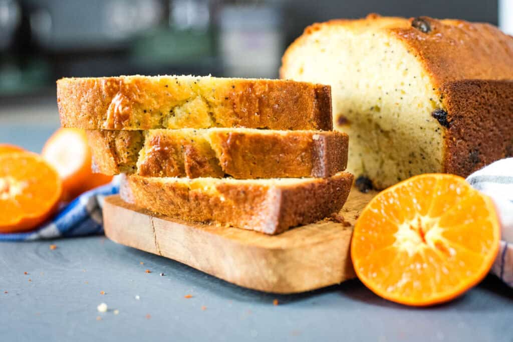 Sliced loaf of bread on a wooden board with two orange slices and a blue-striped cloth nearby on a gray surface.
