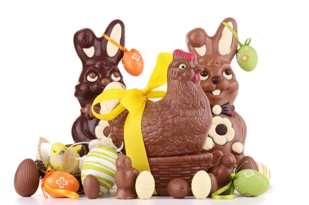 Assorted chocolate Easter bunnies, a chocolate hen with a yellow ribbon, eggs, and small chick figurines arranged on a white background.