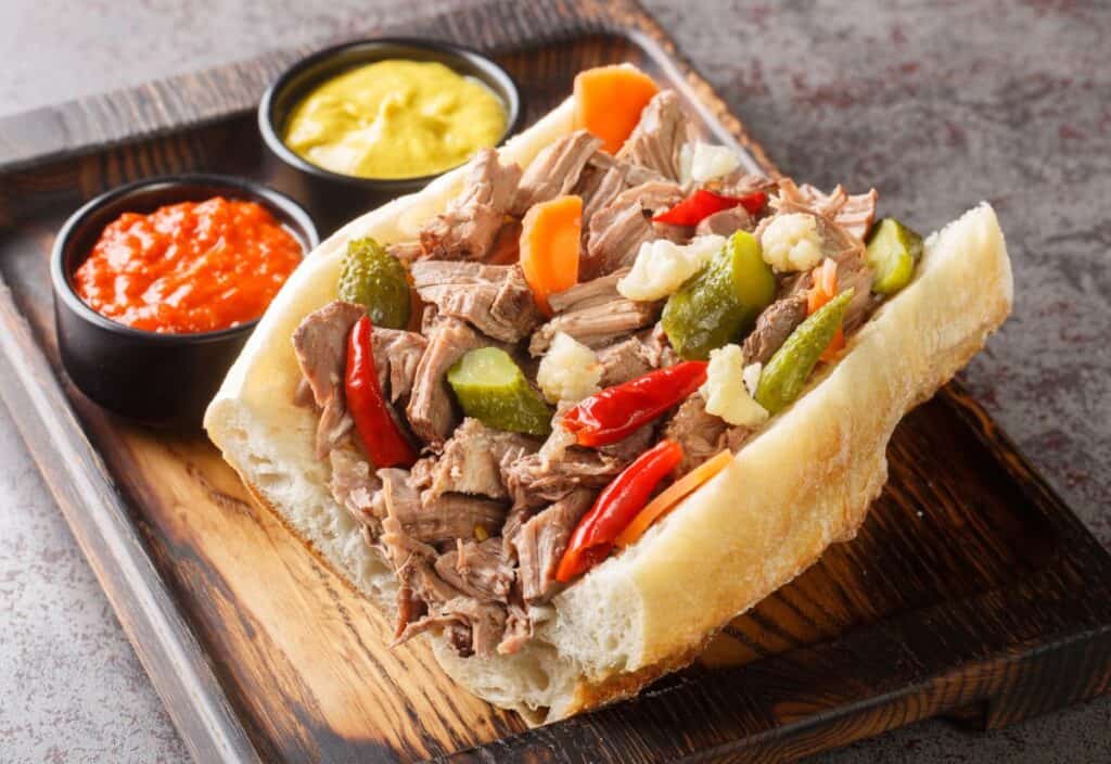 Enjoy Italian beef sandwiches piled high with shredded meat, pickled vegetables, and peppers on a wooden tray, served alongside mustard and red sauce in small bowls.