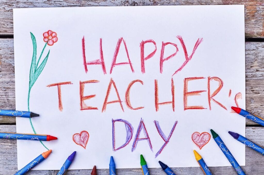 A white paper with "Happy Teacher's Day" written in crayon, decorated with hearts, a flower, and surrounded by colorful crayons on a wooden surface.