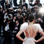 A woman in a white evening gown poses with her back to the camera as a crowd of photographers takes pictures on a red carpet.