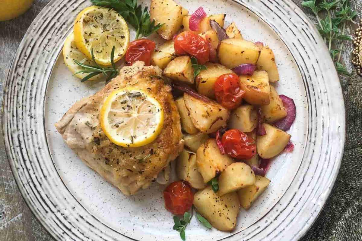 Plate with roasted chicken topped with lemon slices, served alongside roasted potatoes, cherry tomatoes, and red onions, garnished with herbs.