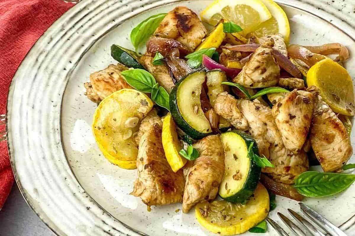 A plate of grilled chicken pieces, sliced zucchini, and yellow squash garnished with basil leaves and lemon wedges. The plate rests on a red cloth with a fork beside it.