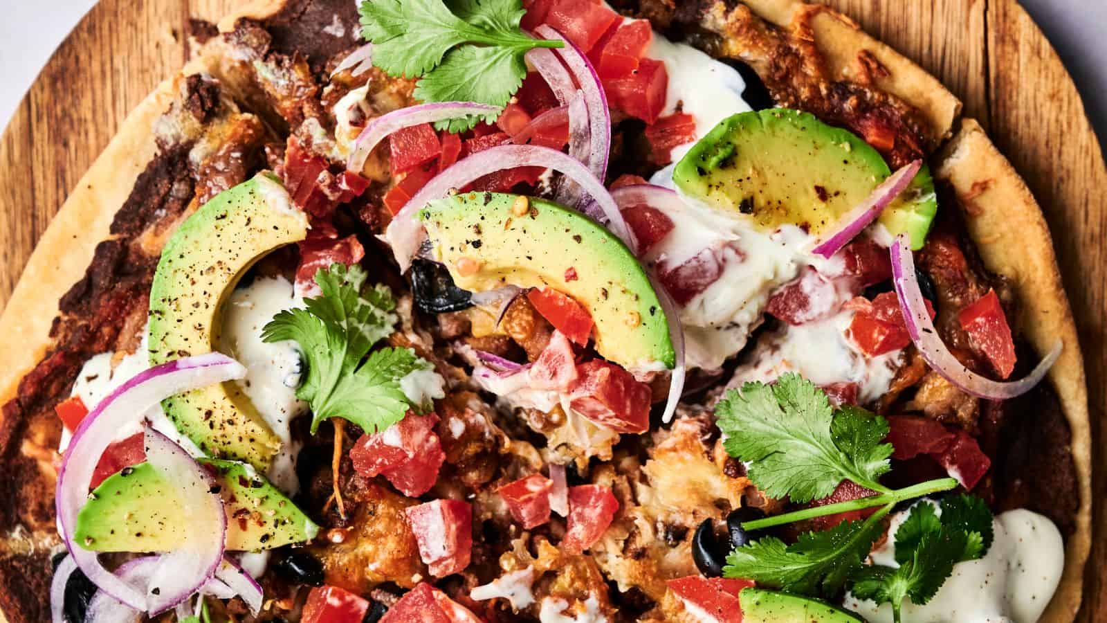 Close-up of a pizza topped with sliced avocado, red onion, tomato, black olives, cilantro, and drizzled sauce on a wooden board.
