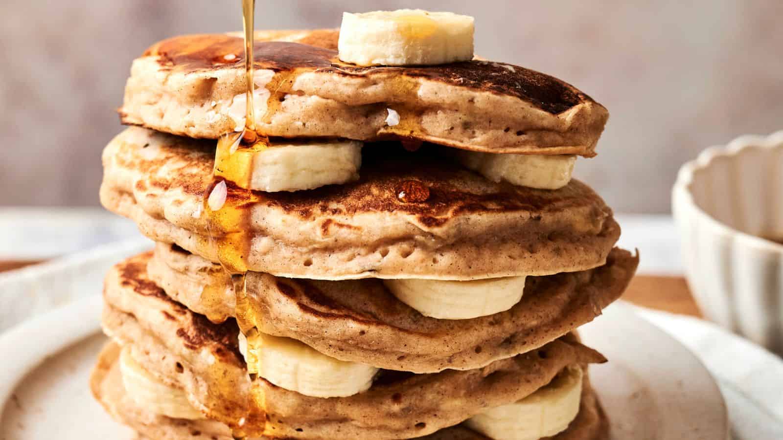 A stack of pancakes with banana slices between layers, topped with more banana and being drizzled with syrup.