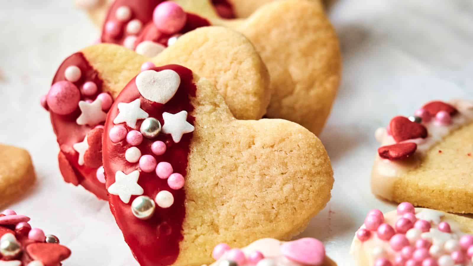 Heart-shaped cookies partially dipped in red icing, decorated with pink, white, and silver sprinkles, including stars and hearts, scattered on a white surface.