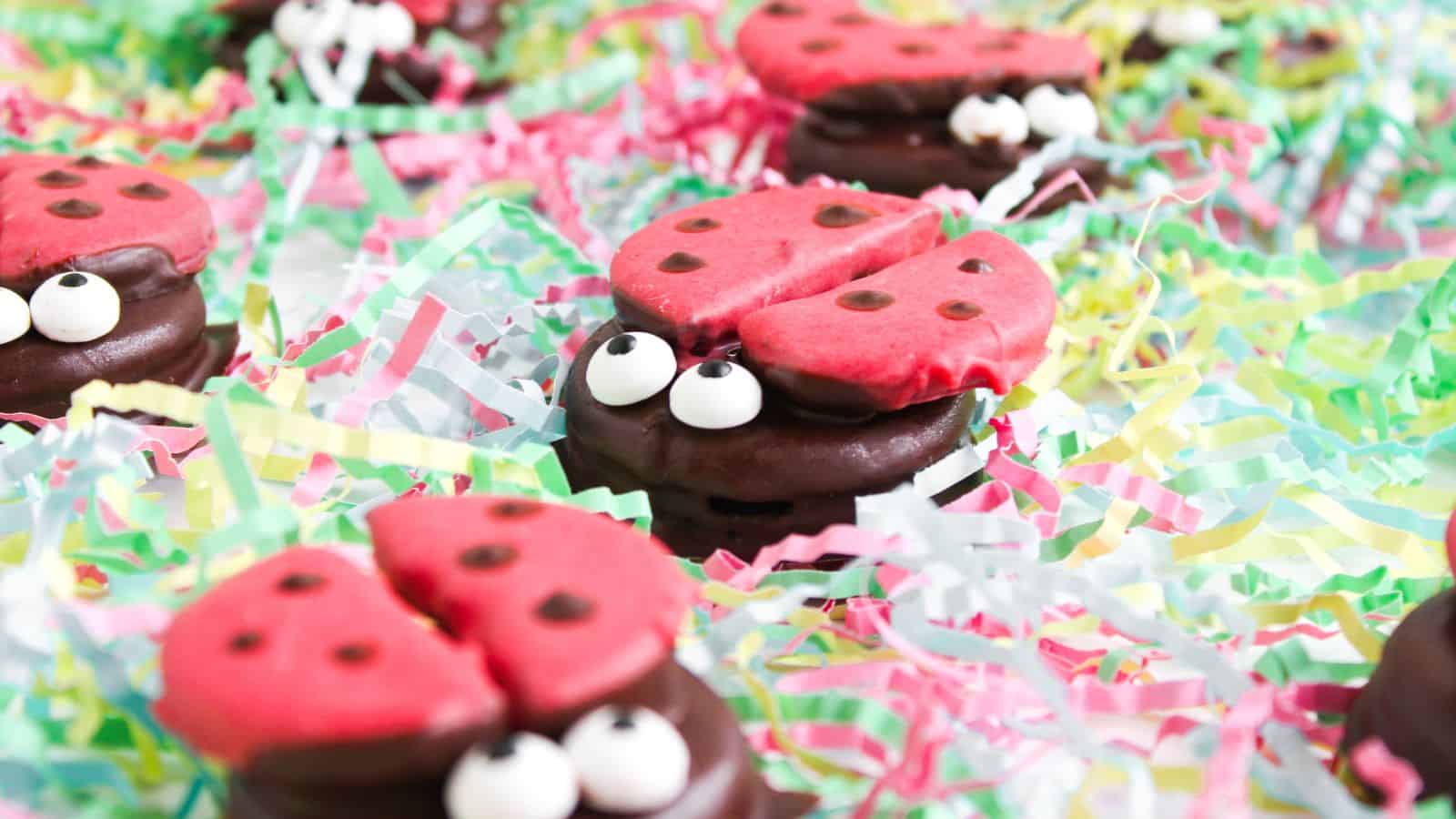 Colorful ladybug-themed cookies with red tops and chocolate bases, decorated with googly eyes, sit on a bed of rainbow paper shreds.