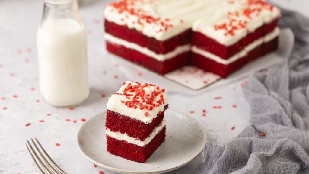 Slice of red velvet cake with cream cheese frosting on a plate, a fork beside it. Whole cake and a milk bottle in the background. Light gray surface and cloth.