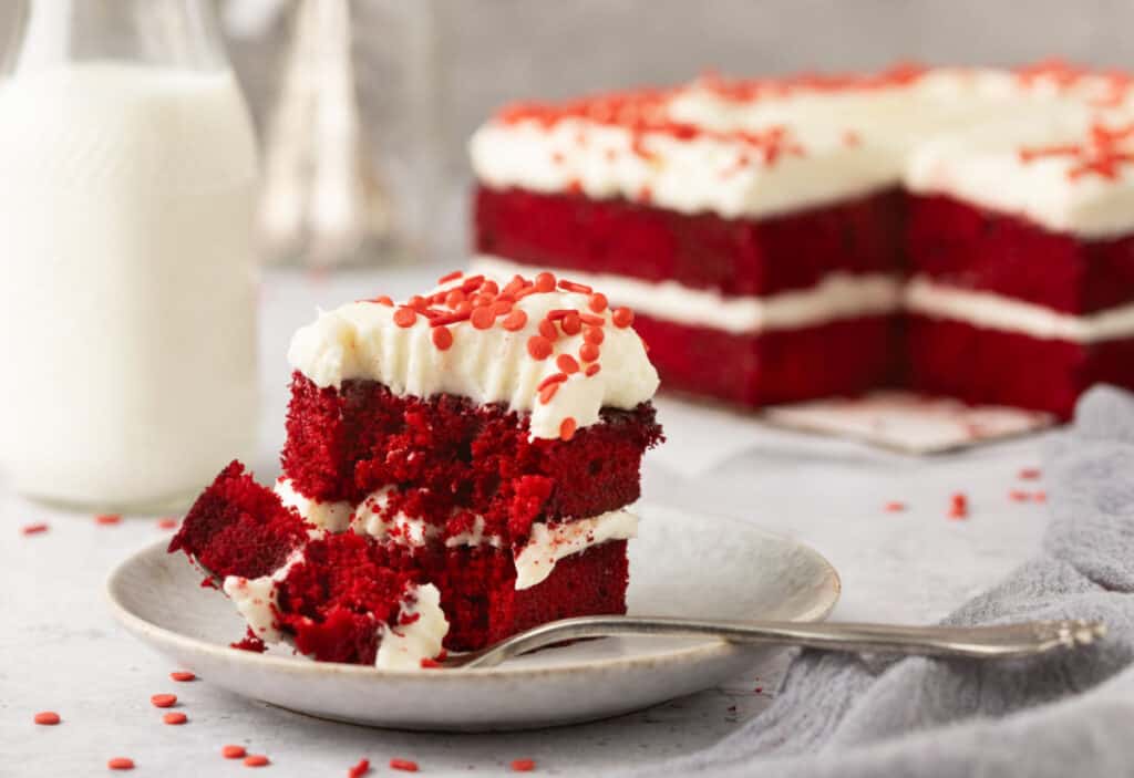 A slice of red velvet cake with cream cheese frosting and red sprinkles on a plate, next to a fork, with a bottle of milk and the rest of the cake in the background.
